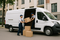 A clean, realistic photo of a modern white cargo van parked on a city street, 
two professional movers in casual work uniforms carefully loading cardboard boxes, 
bright daylight, urban residential background, minimalist commercial style, 
sharp focus, high resolution, friendly and trustworthy atmosphere.